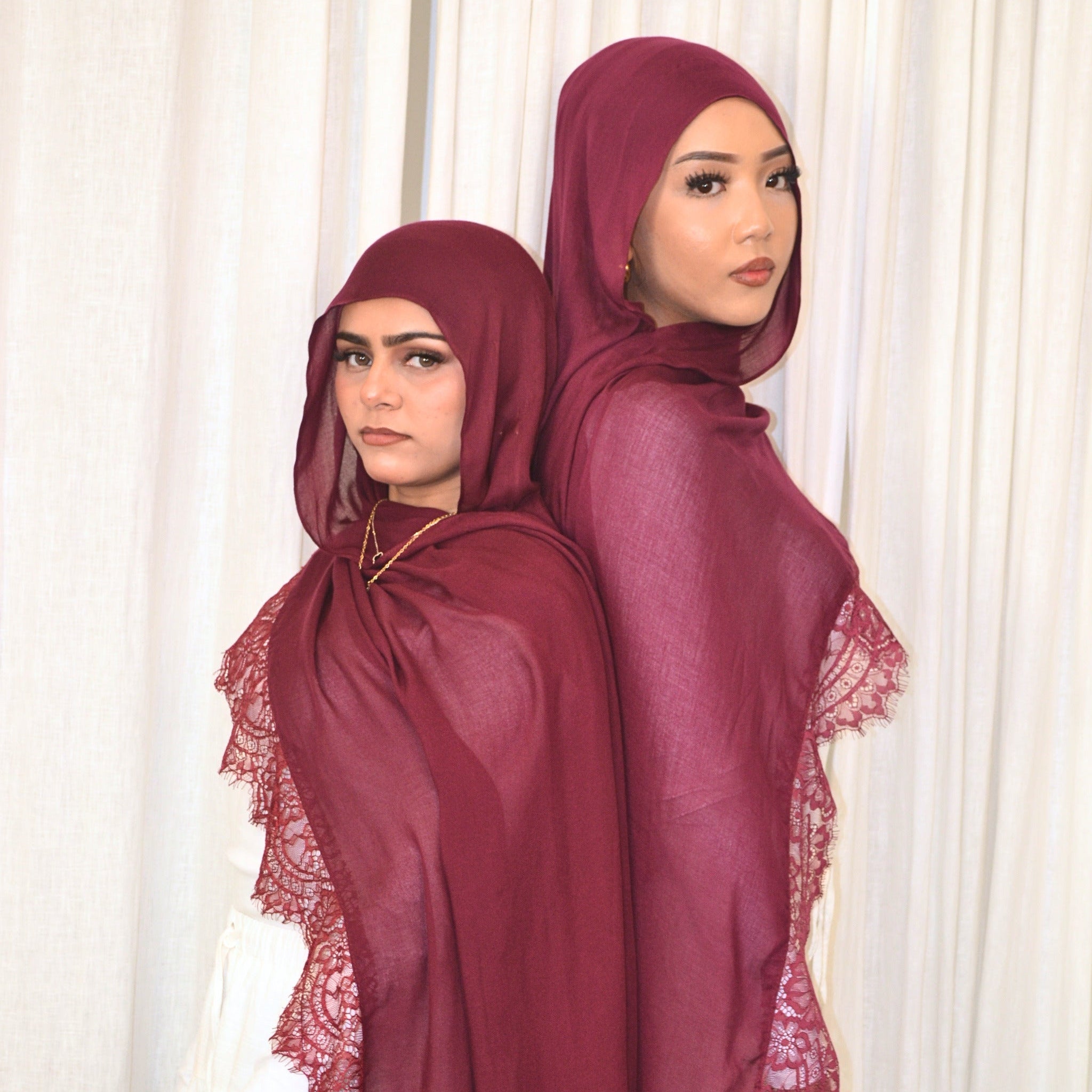 Two women modeling matching deep burgundy hijabs and shawls with lace trim, posed back-to-back in front of a white curtain backdrop.